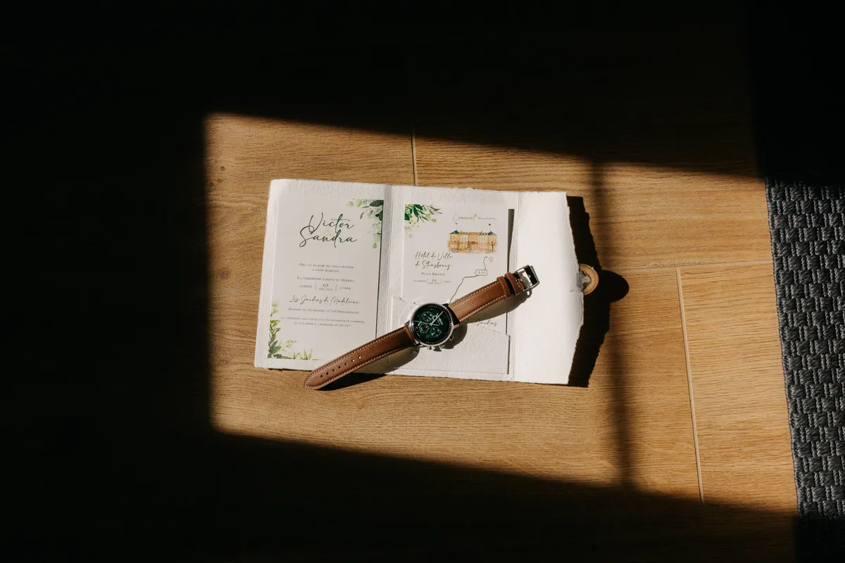 Invitación de boda y reloj del novio sobre el suelo de madera, con la luz de la mañana entrando de lado
