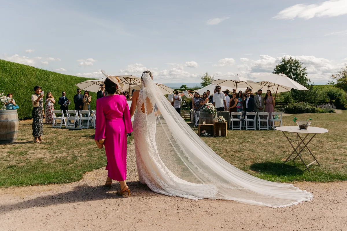 Sandra entrando a la ceremonia con la cola del vestido y el velo largo sobre el camino de grava