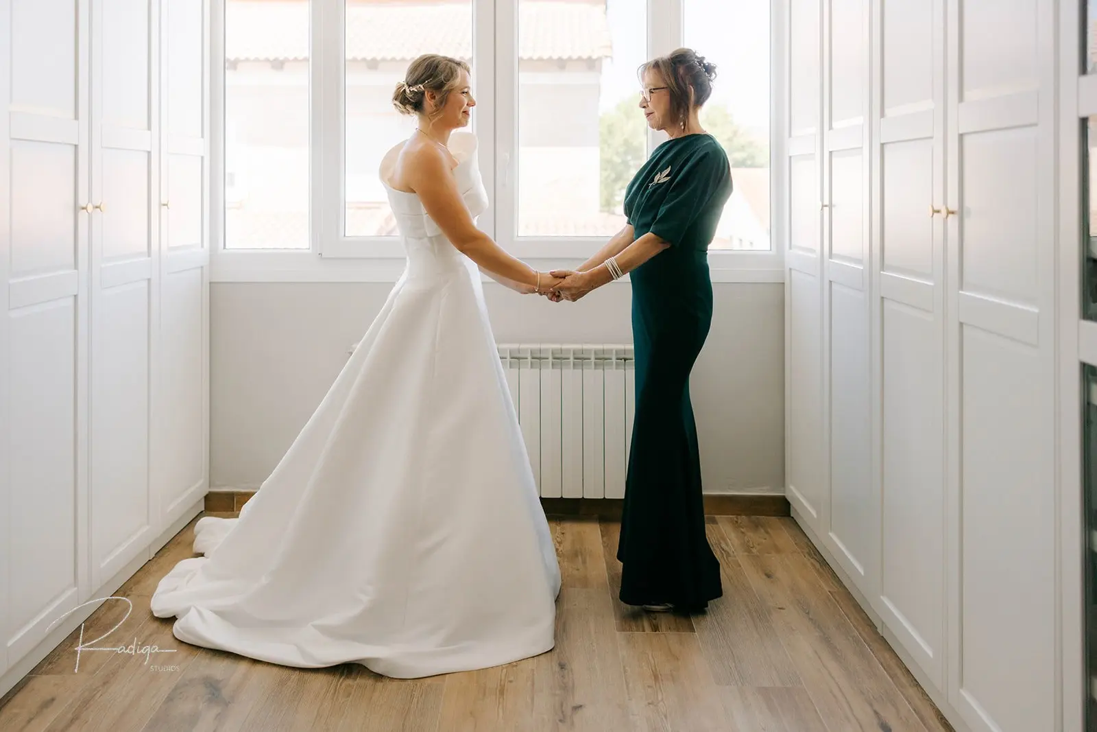 La novia, ya vestida, de la mano de su madre junto a la ventana