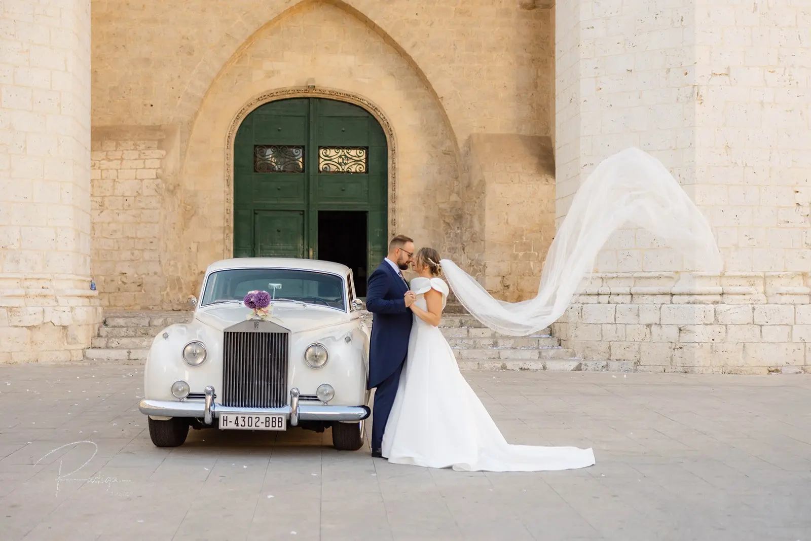 Los novios ante la torre-pórtico de San Benito el Real con el coche clásico y el velo al viento