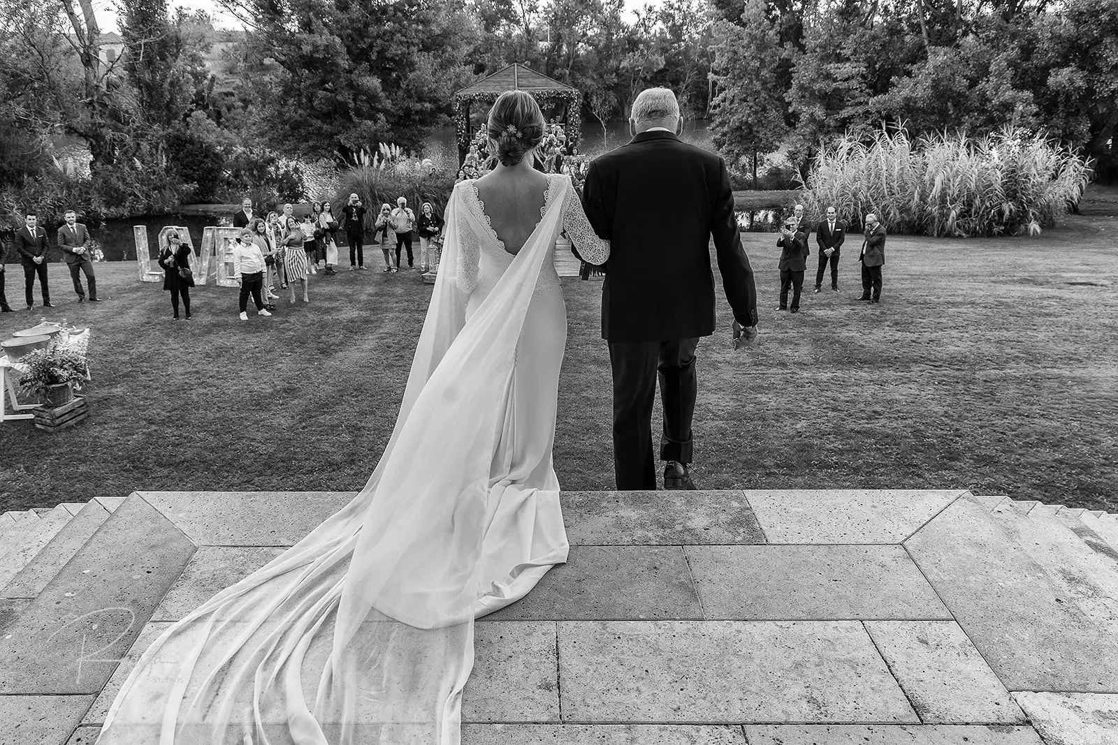 La novia camina hacia la ceremonia del brazo del padrino, con el velo sobre la hierba, en blanco y negro