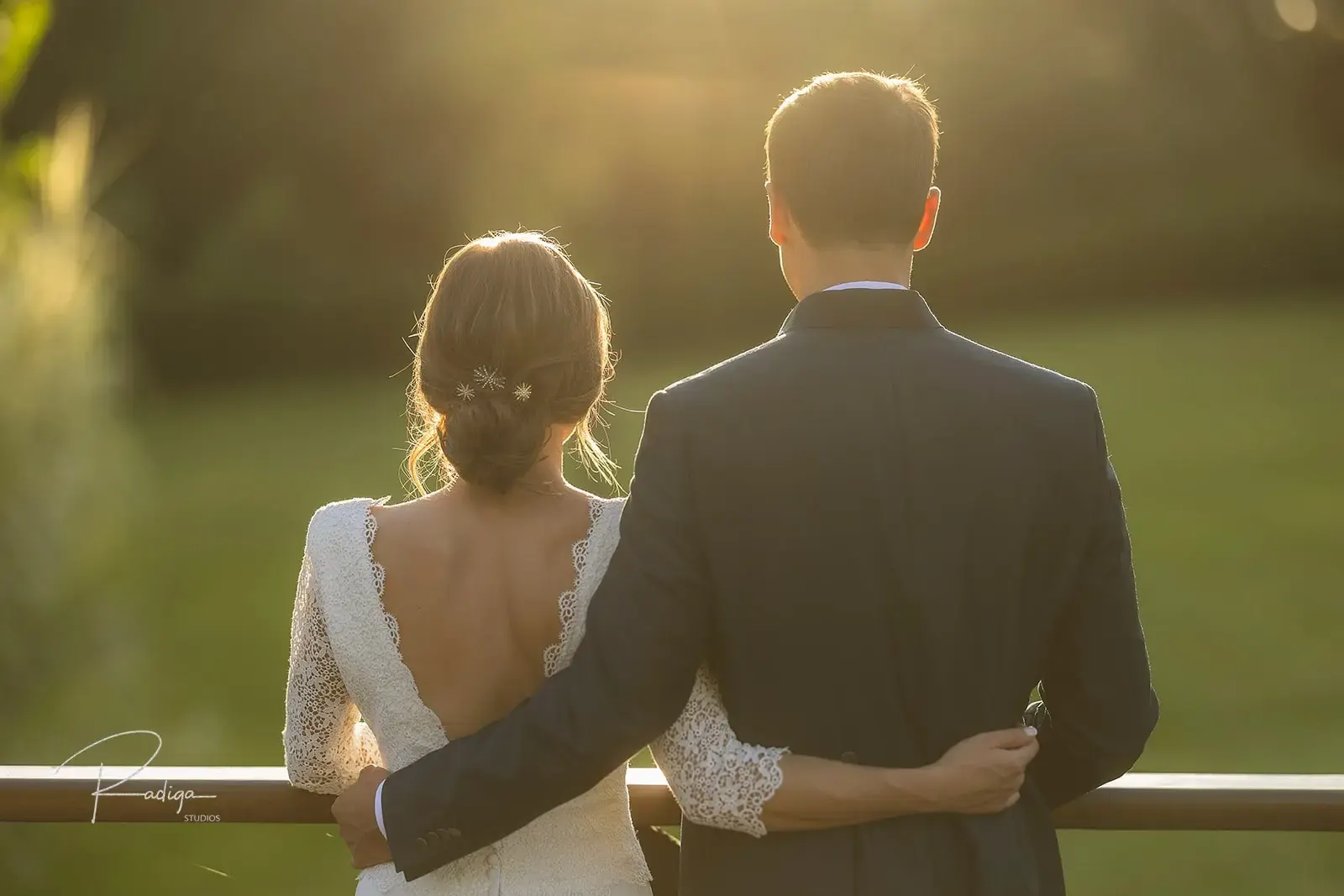 Los novios de espaldas, abrazados junto a la barandilla, mirando el jardín al atardecer