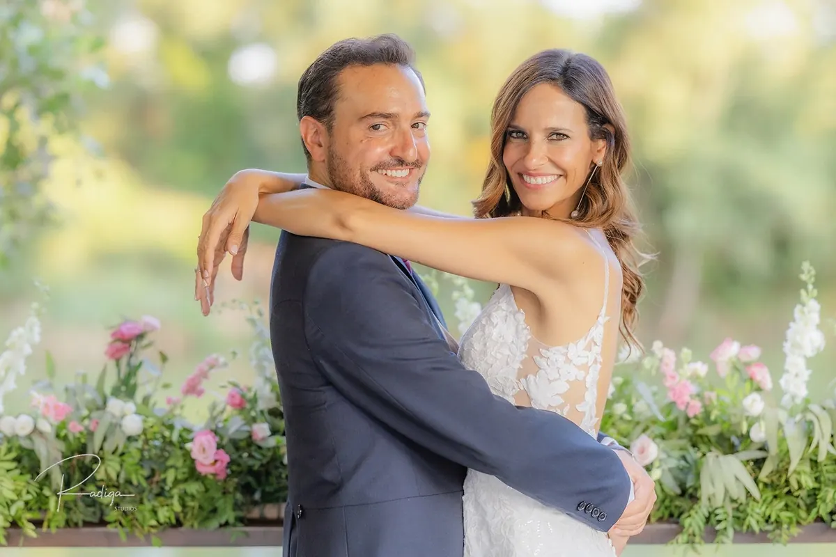 Los novios abrazados y sonrientes frente a las flores del jardín del AC Palacio de Santa Ana