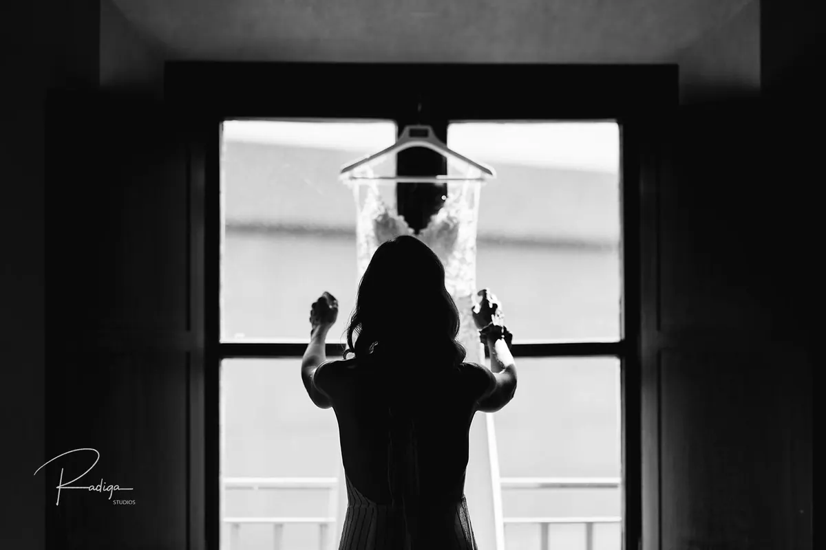 Silueta de la novia frente a la ventana con el vestido colgado, en blanco y negro