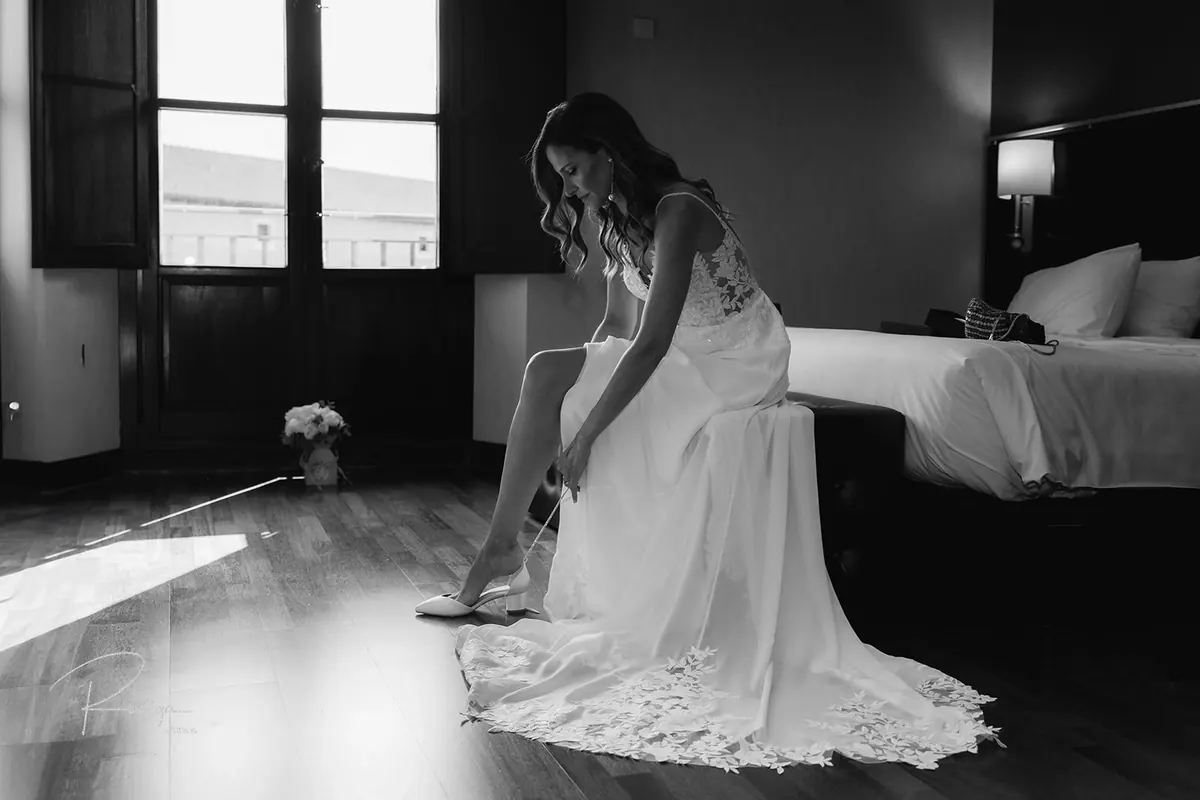 La novia se calza sentada en la cama, en blanco y negro