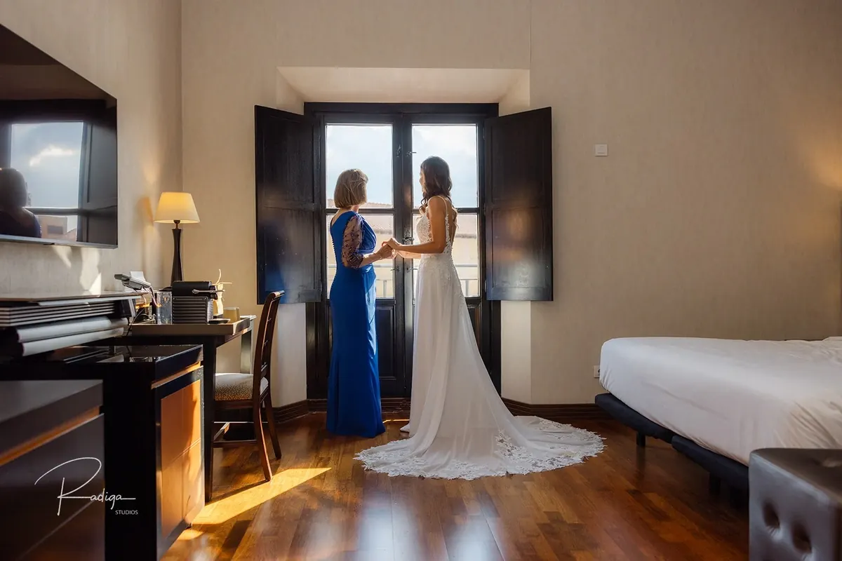 La novia, ya vestida, junto a la ventana con una invitada de azul
