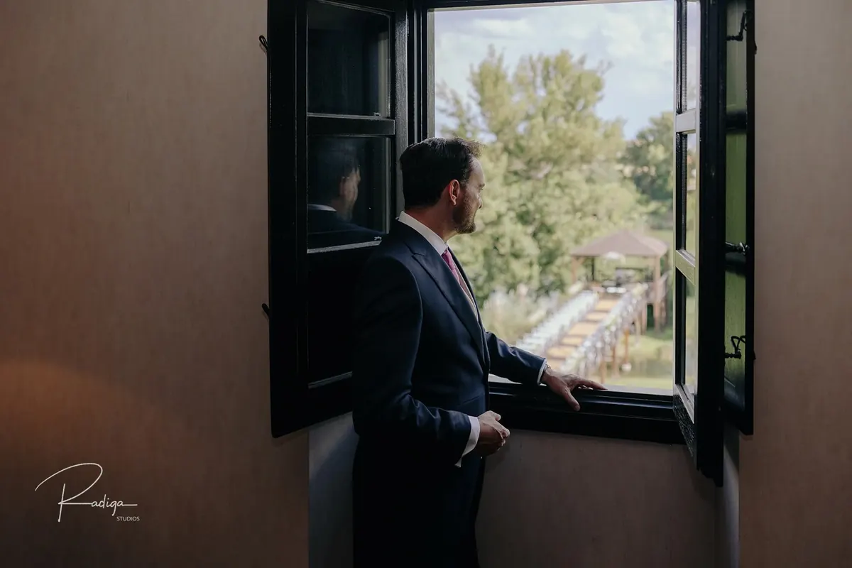 El novio, ya vestido, mira por la ventana hacia los jardines