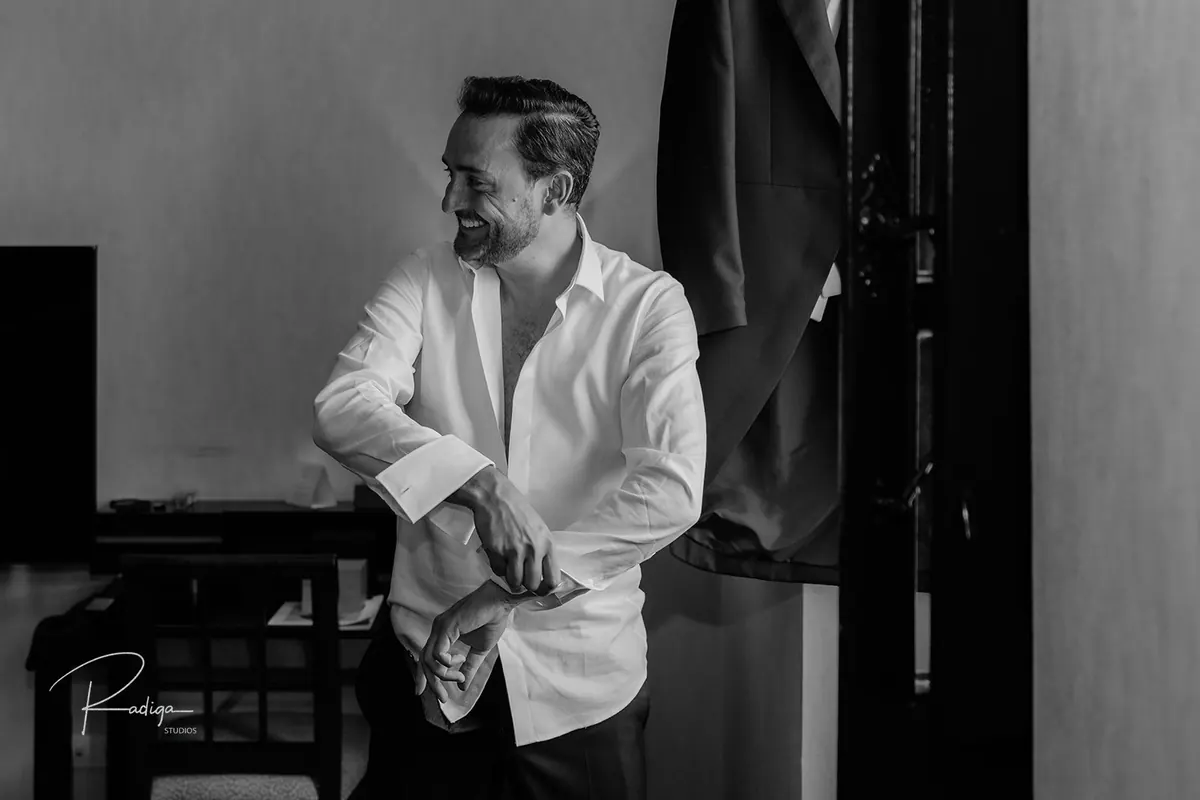El novio sonríe apoyado en la ventana con la camisa blanca, en blanco y negro