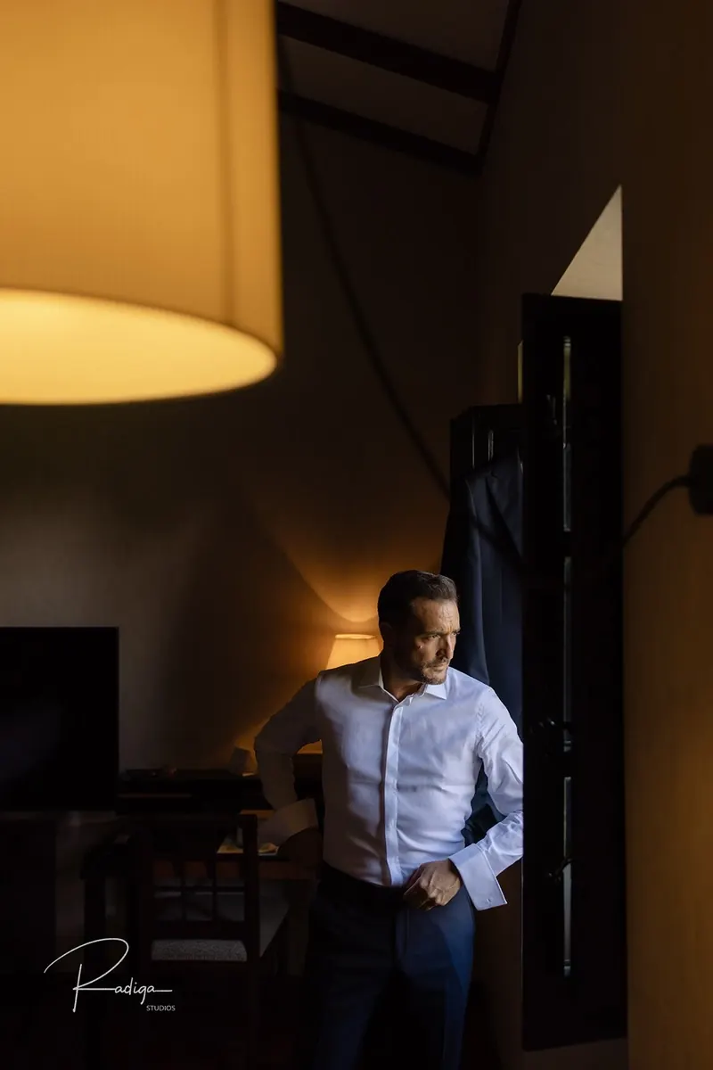 El novio se abrocha la camisa junto a la ventana de la habitación en penumbra