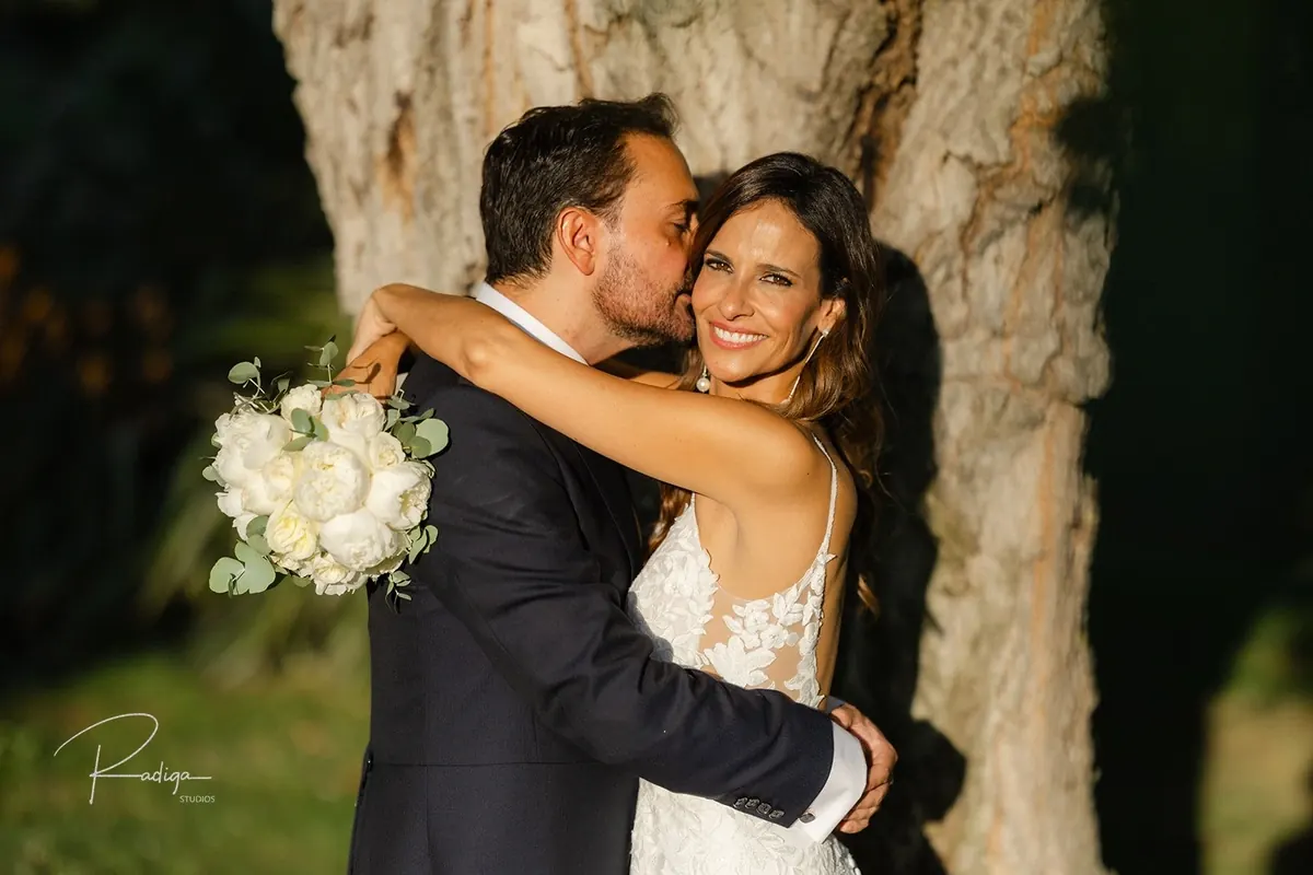 Los novios abrazados junto al tronco de un gran árbol del jardín
