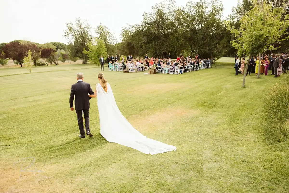 La novia avanza por el césped hacia la ceremonia con el velo largo detrás