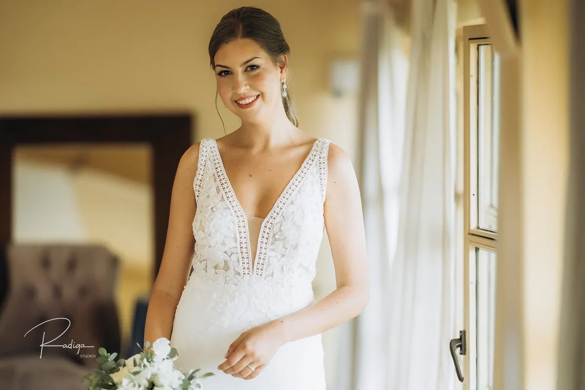 La novia sonríe, ya vestida, junto a la ventana con el ramo