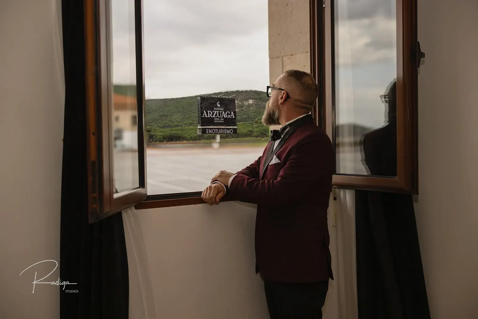 The groom in a burgundy tuxedo looking out of the hotel window at Arzuaga