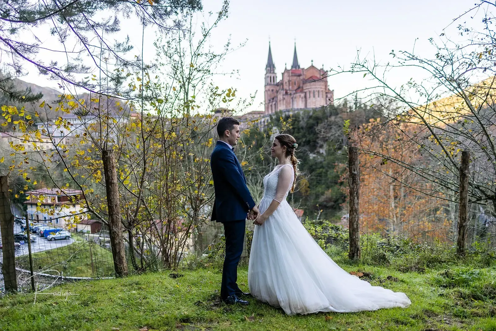 Los novios junto a un muro de piedra con la Basílica de Covadonga al fondo