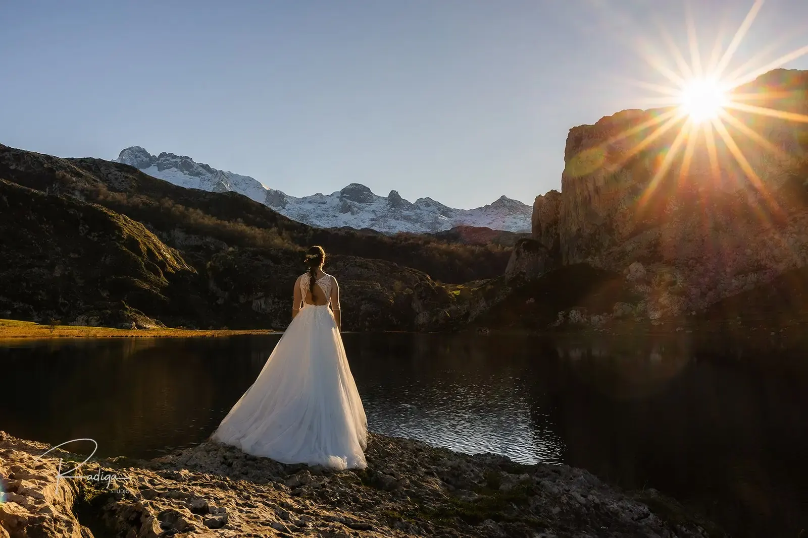 La novia sola a contraluz a orillas del lago, con el sol asomando tras la montaña