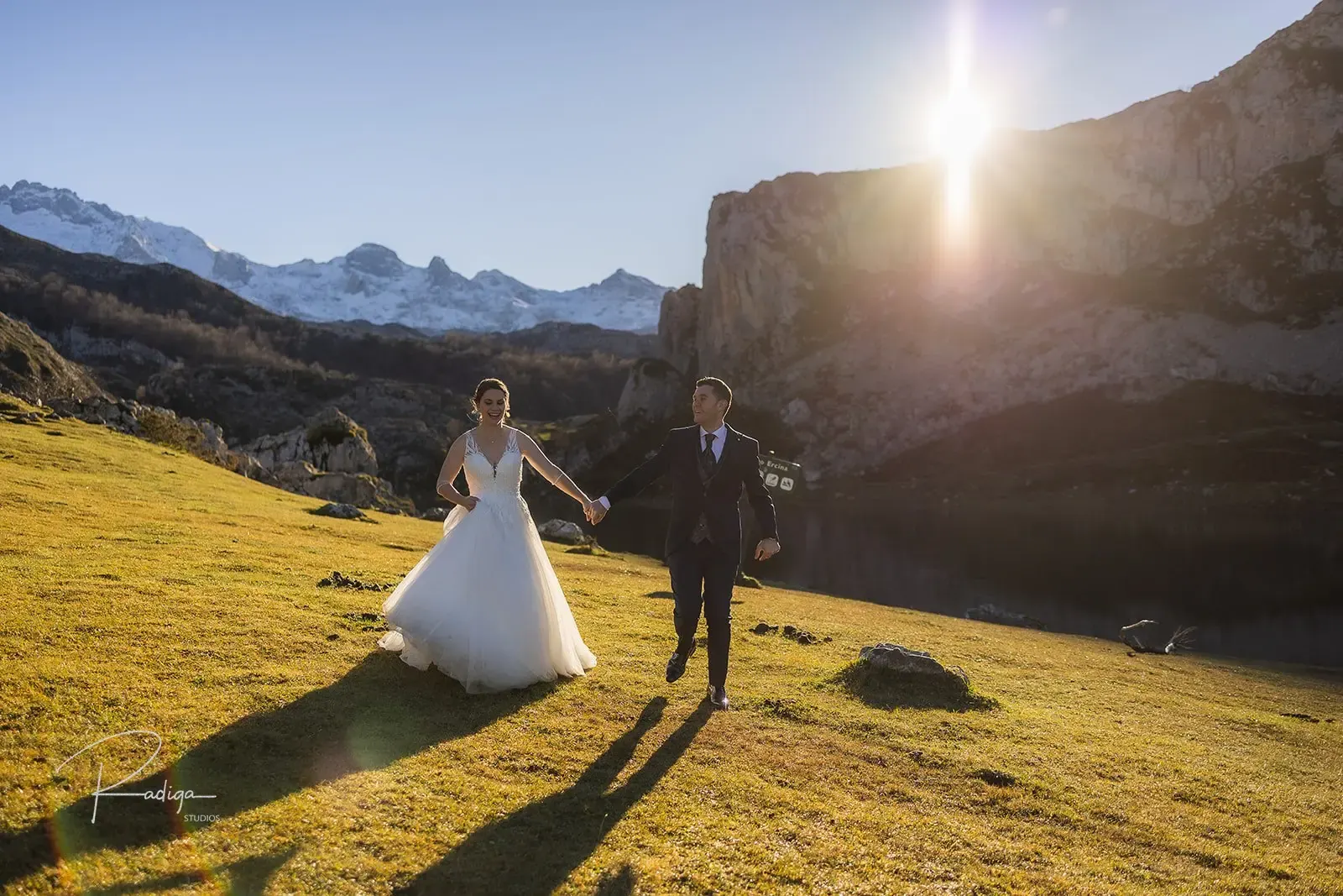 Los novios caminan por la hierba con el sol de frente y los picos nevados detrás