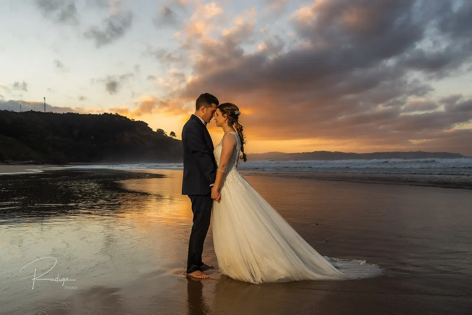 Los novios abrazados en la playa bajo un cielo de nubes al atardecer