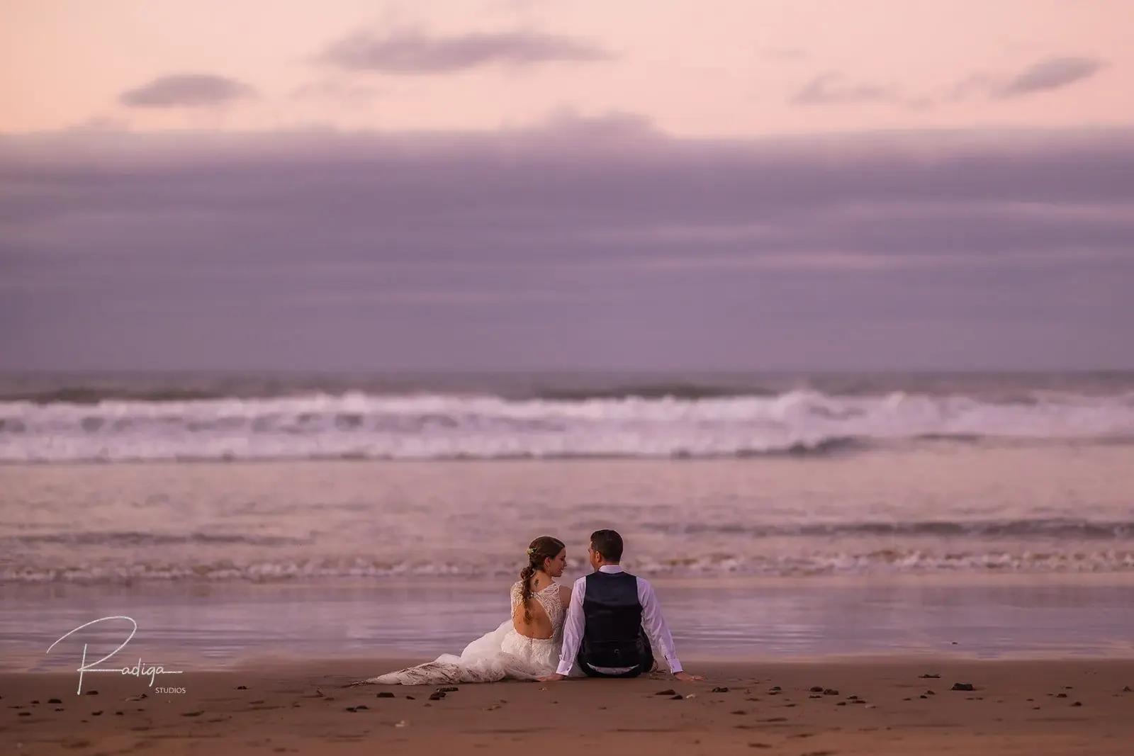 Los novios sentados en la arena frente al mar con el cielo rosado del final del día