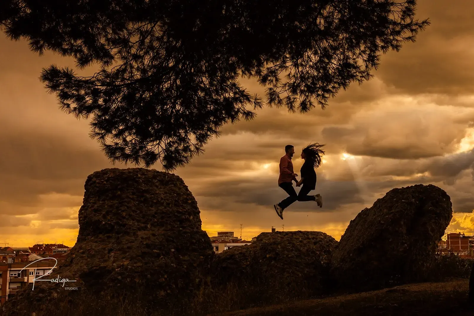 La pareja salta entre dos grandes rocas bajo un árbol, a contraluz del atardecer