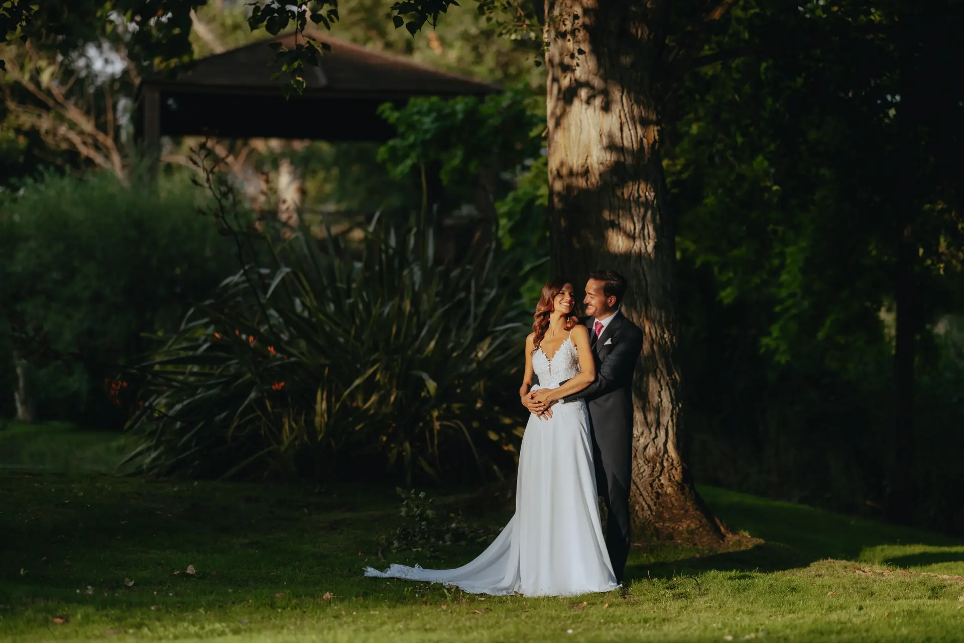 Los novios abrazados bajo un gran árbol del jardín de la finca, con la última luz de la tarde