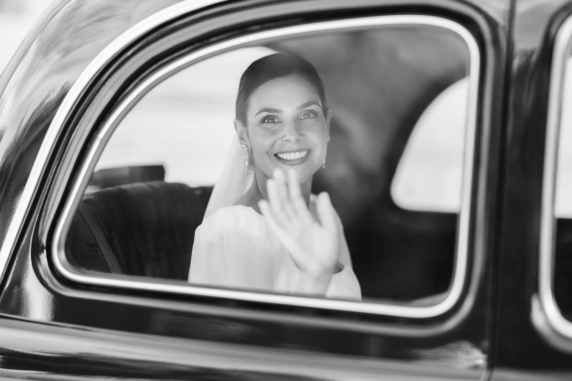 La novia saluda sonriente desde la ventanilla del coche camino de la ceremonia, en blanco y negro