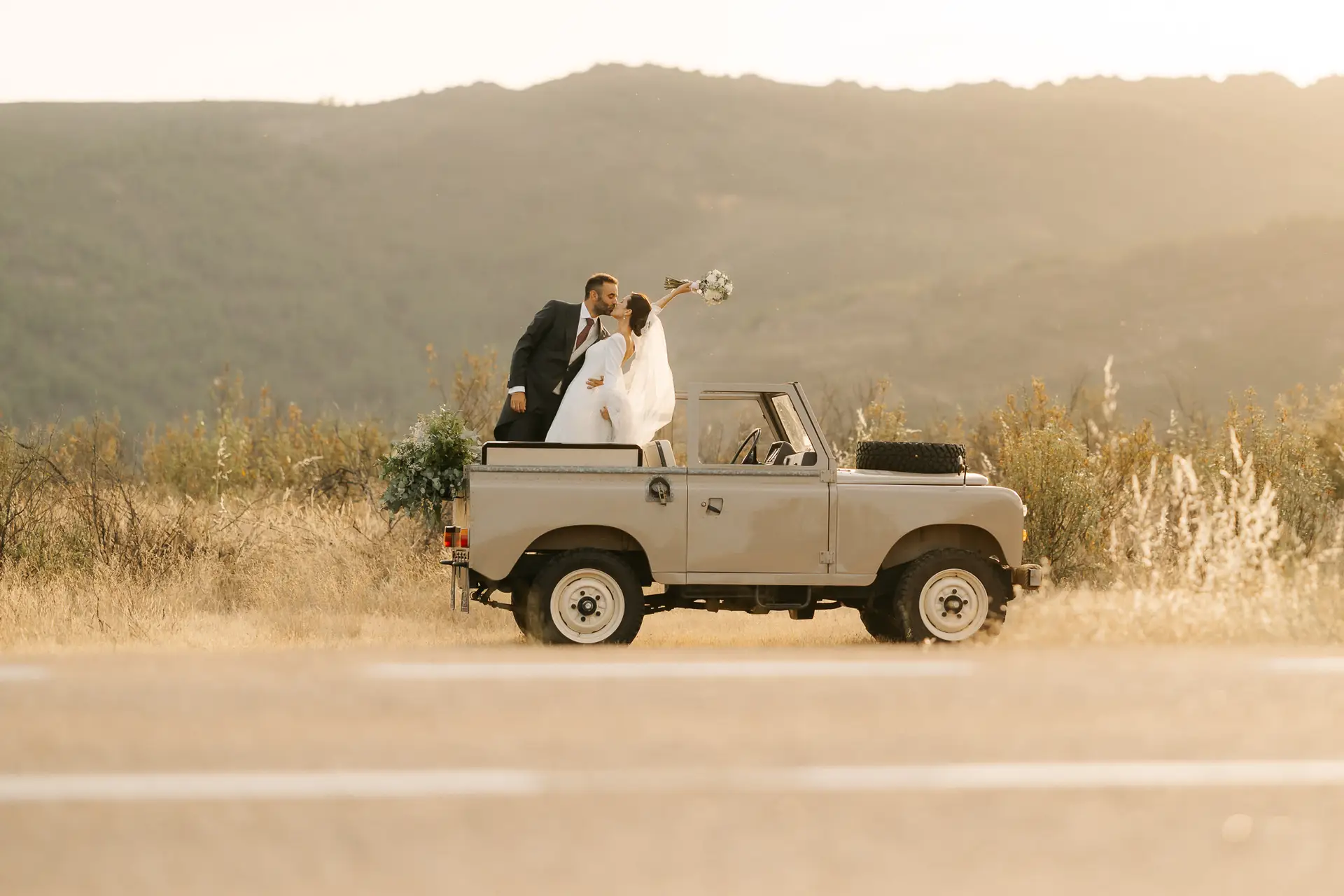 Los novios se besan de pie en la parte trasera de un coche clásico descapotado, con el ramo en alto y el campo al atardecer
