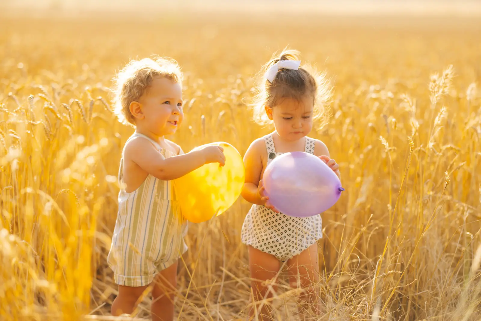 Dos niños pequeños con un globo amarillo y otro morado en un campo de trigo al atardecer