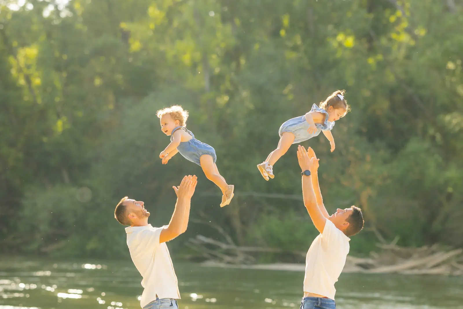 Dos padres lanzando por el aire a sus hijos pequeños junto al río, a contraluz